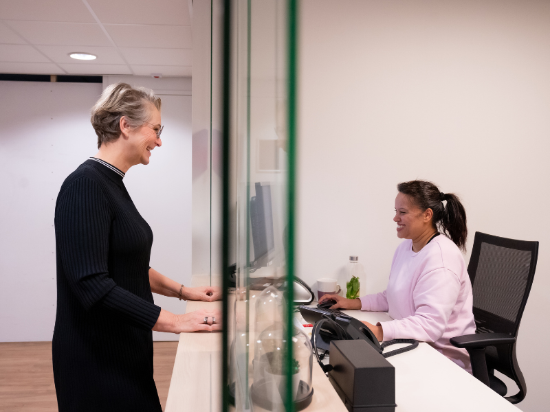Vrouw in gesprek aan balie bij huisartsenpraktijk met de doktersassistente.